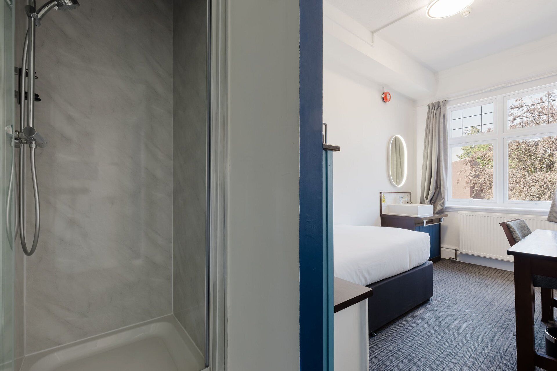 A hotel room with a shower, a bed, a desk, a window with a view, and a mirror.