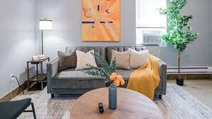 A modern living room with a grey sofa, a round coffee table with a vase of flowers, a floor lamp, and a large abstract painting on the wall. - Lancaster Art Suite 2 - 4BR near Drexel & Penn (Philadelphia)