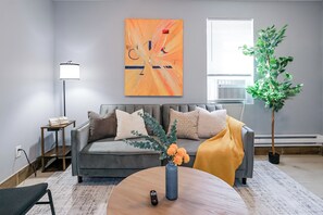 Una sala moderna con un sofá gris, una mesa de centro redonda con un jarrón de flores, una lámpara de pie y un cuadro abstracto grande en la pared.