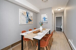 Comedor con una larga mesa dispuesta para seis personas, sillas anaranjadas, un jarrón blanco con flores y arte abstracto en las paredes.