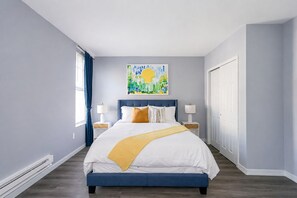 Un dormitorio con una cama, dos mesitas de noche, una ventana con cortinas, un cuadro en la pared y una puerta.