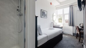 A hotel room with a bed, a shower, a desk with a chair, a window with curtains, and a picture on the wall. - Comfortable Single Room (Watford)