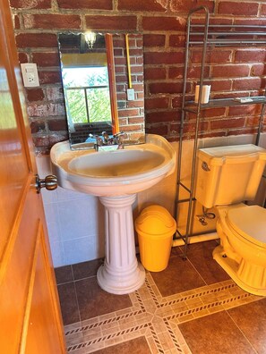 Ein Badezimmer mit einem Waschbecken auf Säule, einer Toilette, einer gelben Mülltonne und einem Metallregal.