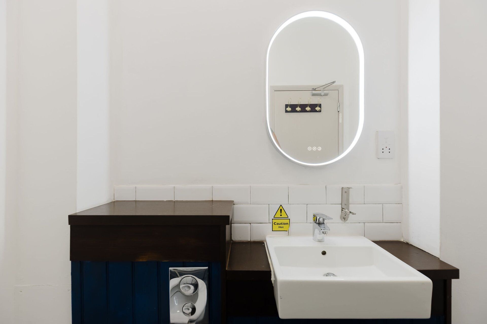 Ein Badezimmer mit weißem Waschbecken, einem Spiegel mit Beleuchtung, einem Warnschild und einem fest montierten Stromkasten.
