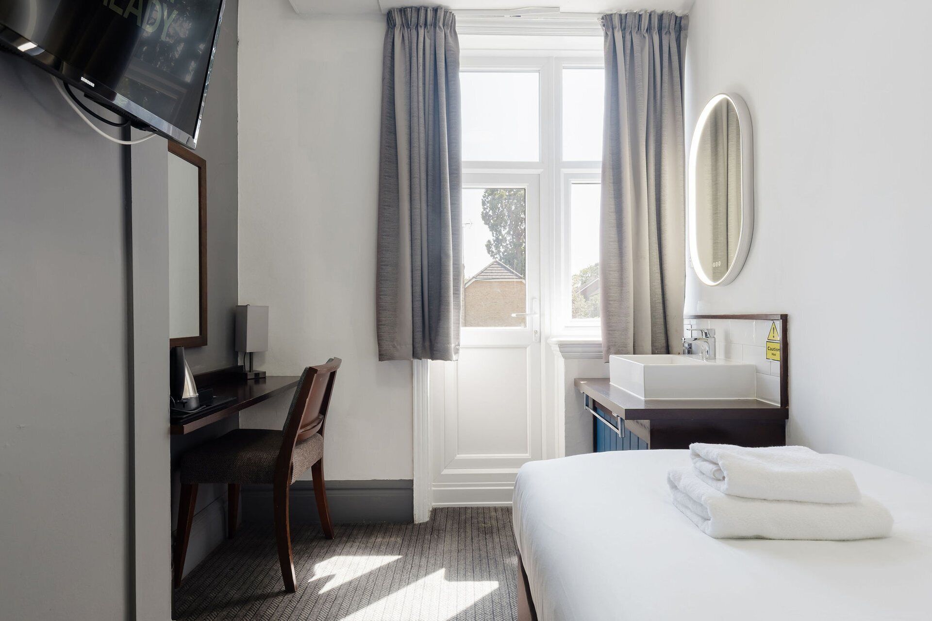 Ein Hotelzimmer mit Bett, Schreibtisch, Stuhl, Fernseher, Fenster mit Vorhängen, Spüle und Spiegel.