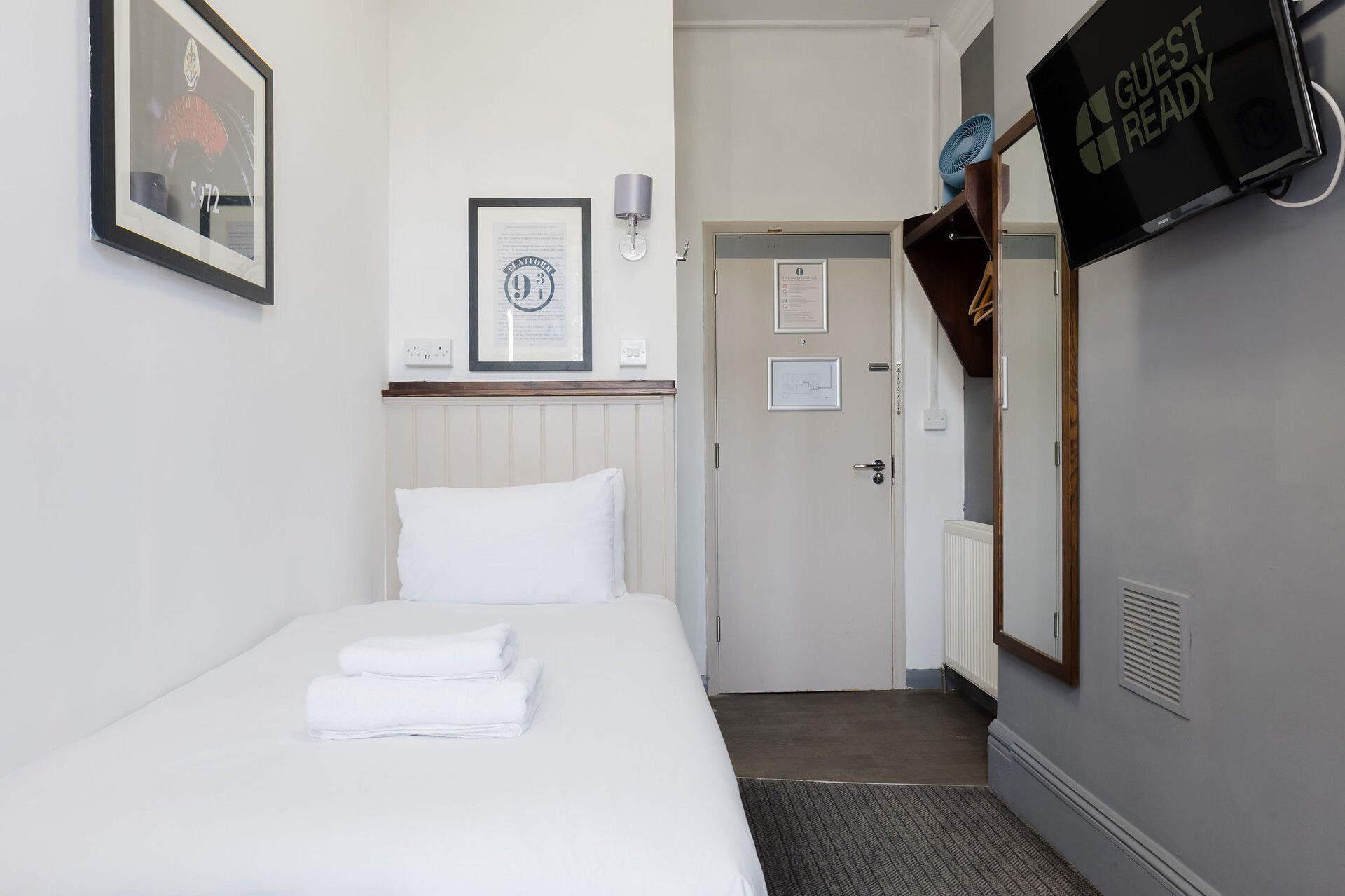 A hotel room with a bed, a framed certificate on the wall, a door, and a TV screen displaying 'GUEST READY'.