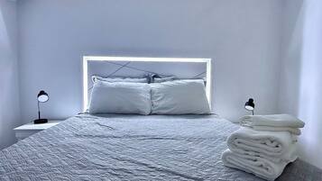 Una cama bien hecha con cabecera, una lámpara de noche y una pila de toallas dobladas.