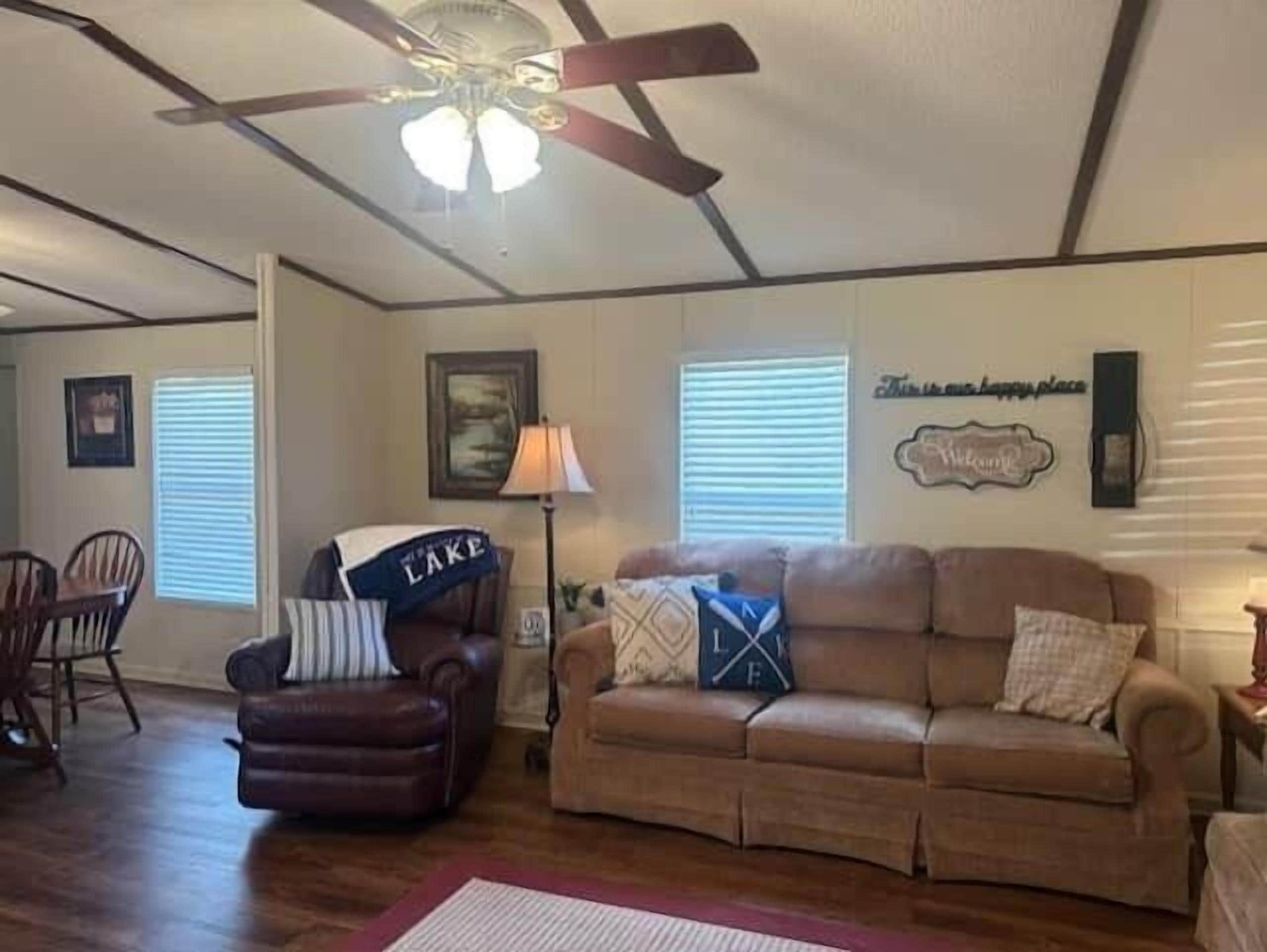 Un salon avec un canapé en cuir marron, un fauteuil marron, un ventilateur de plafond et un mur orné d’une pancarte de bienvenue.