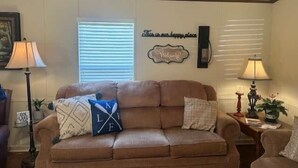 Una sala de estar con un sofá marrón, dos almohadas, un reloj de pared, un letrero de bienvenida enmarcado y una ventana con persianas.
