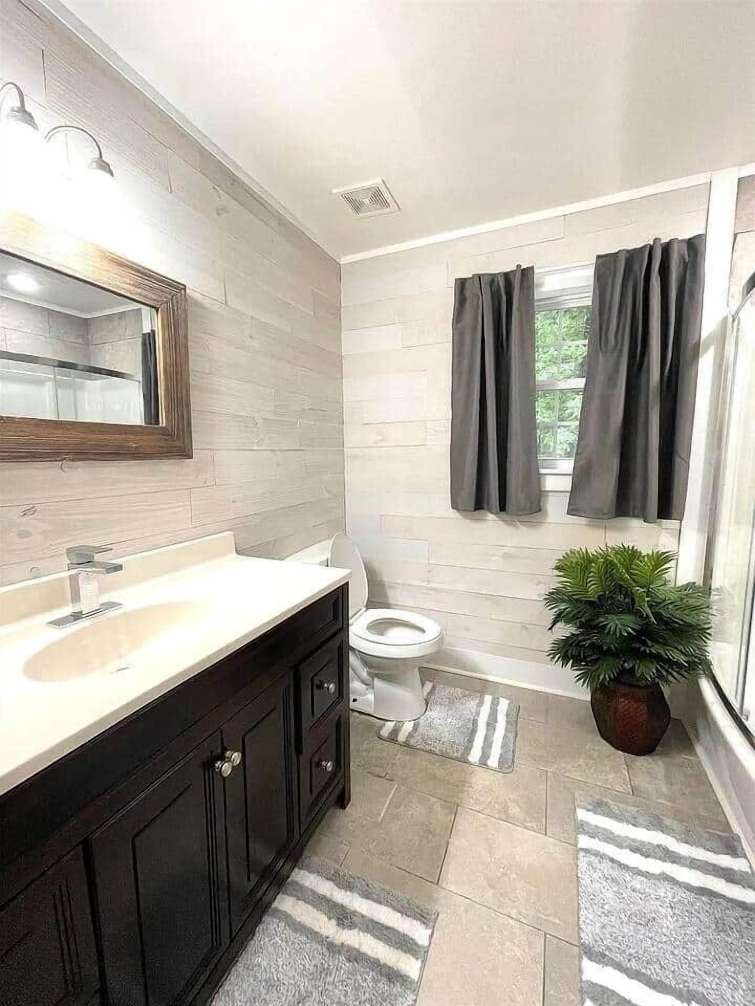 Un baño con un lavamanos blanco, armarios oscuros, un espejo, un inodoro, zona de ducha y una planta en maceta.