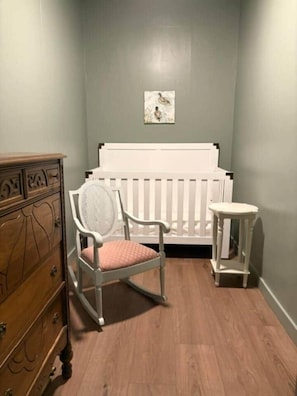 A room with a white crib, a rocking chair, a small stool, and a wooden dresser.