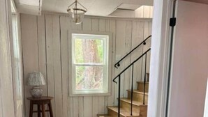 A wooden staircase with a handrail, a window with a view of trees, and a lamp on a stool. - The Bungalow at Bear Creek (Tishomingo County)