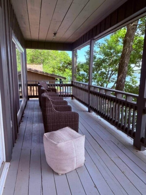 A covered porch with wicker furniture, a wooden deck, and a view of trees and a building in the background.