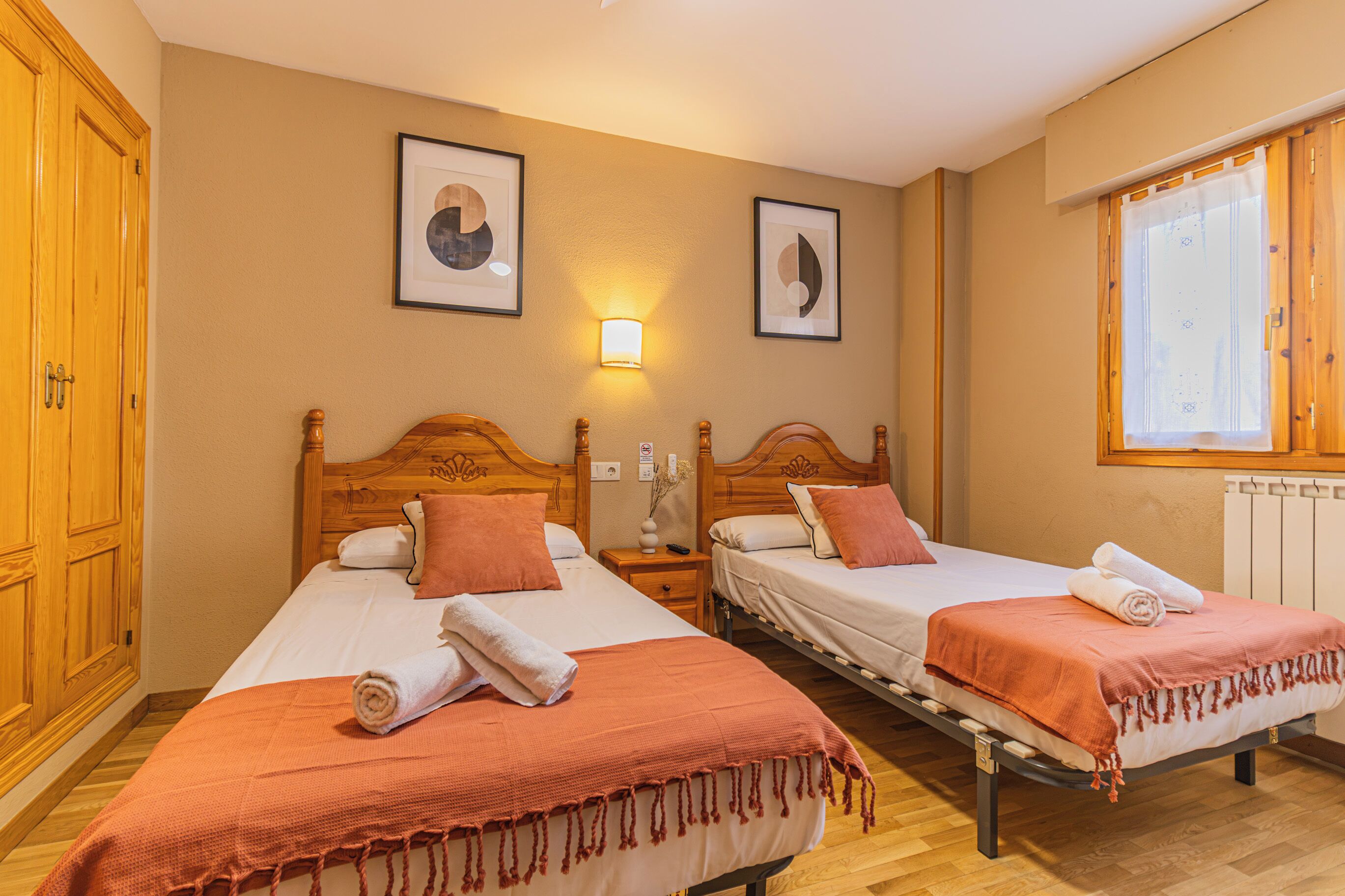Habitación de hotel con dos camas, cabeceros de madera, una mesita de noche, una lámpara de pared y un ventanal con cortinas.