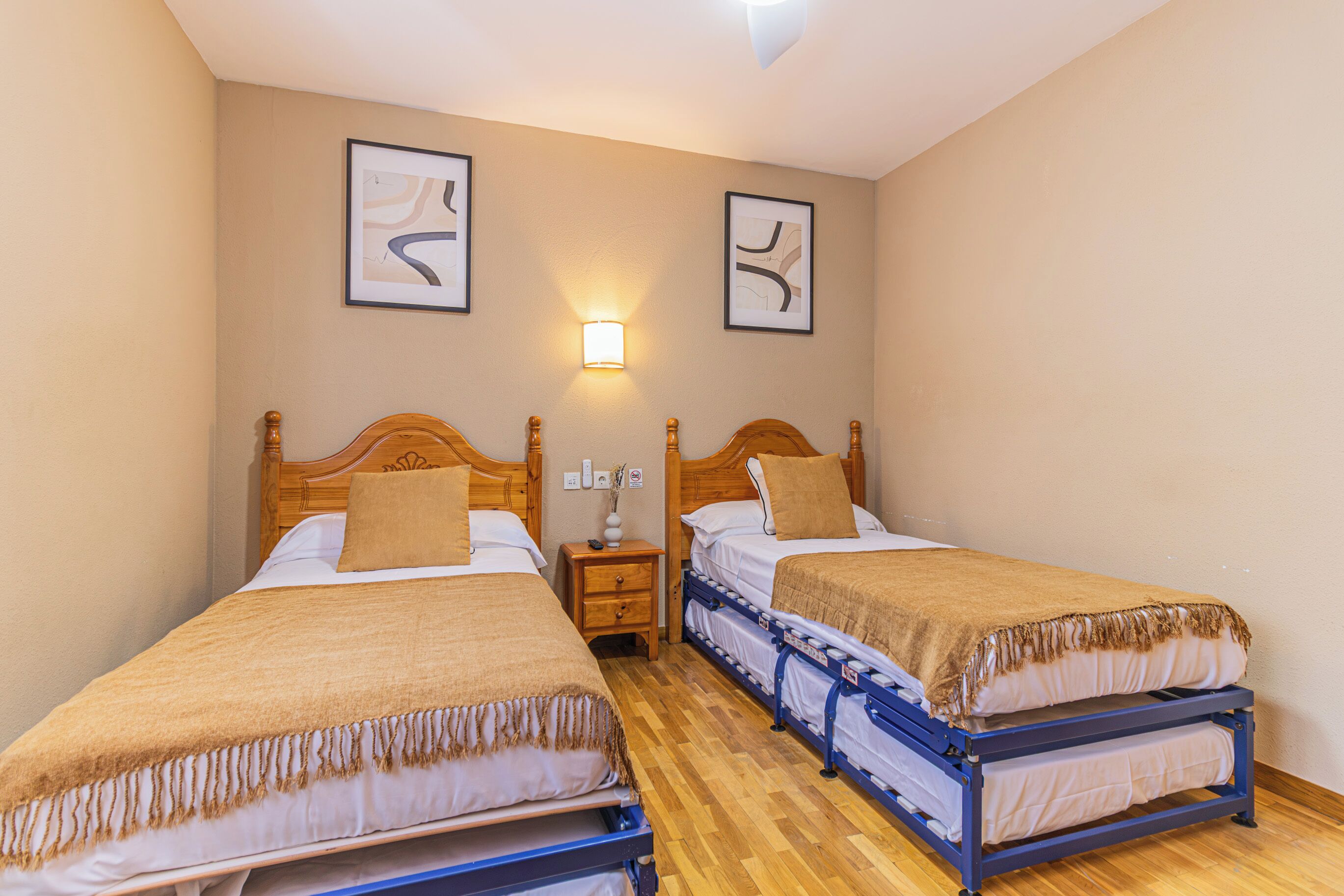 Habitación de hotel con dos camas individuales, una mesita de noche, una lámpara de pared y dos cuadros abstractos enmarcados.