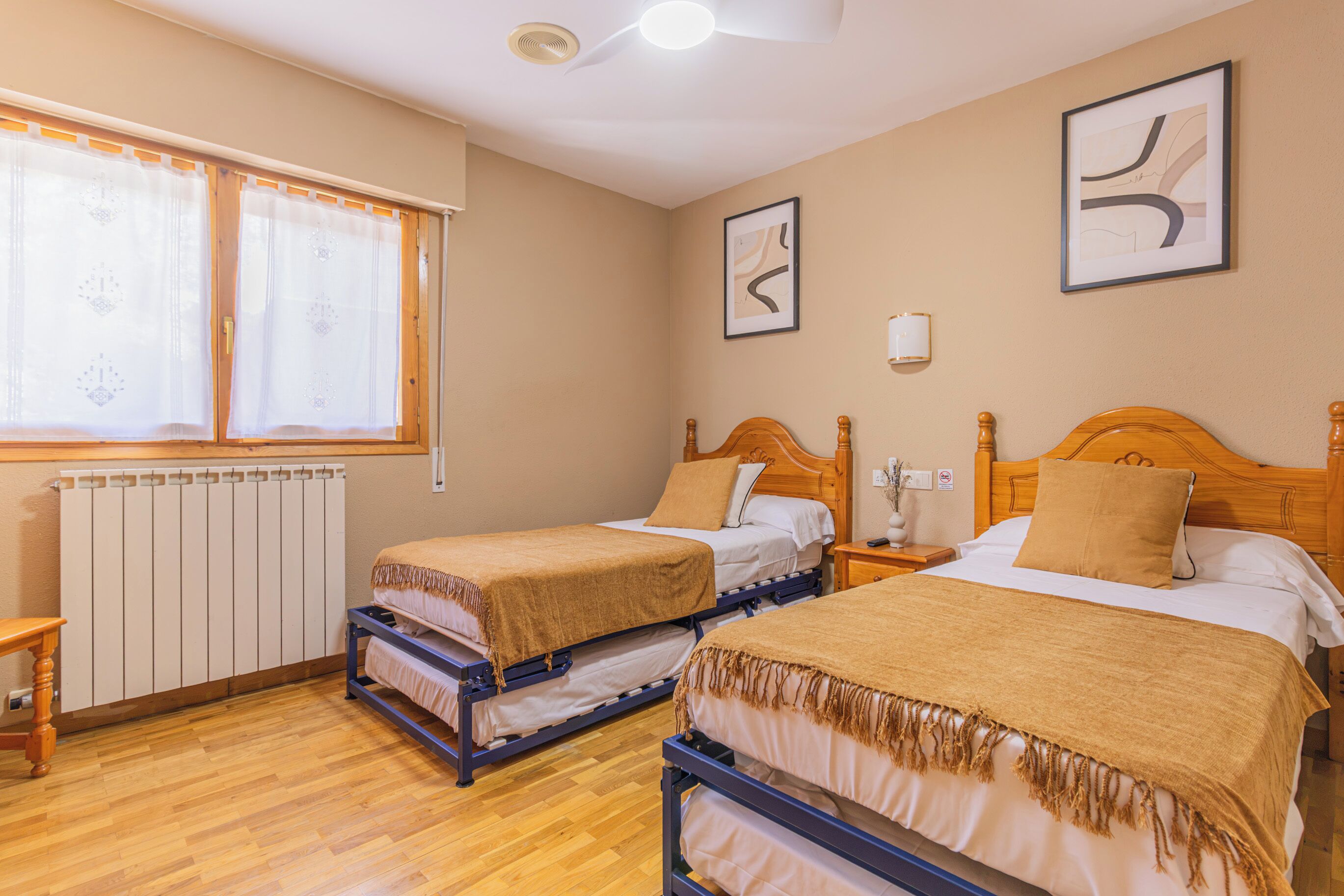 A hotel room with two beds, a radiator, a window with curtains, and a ceiling fan.