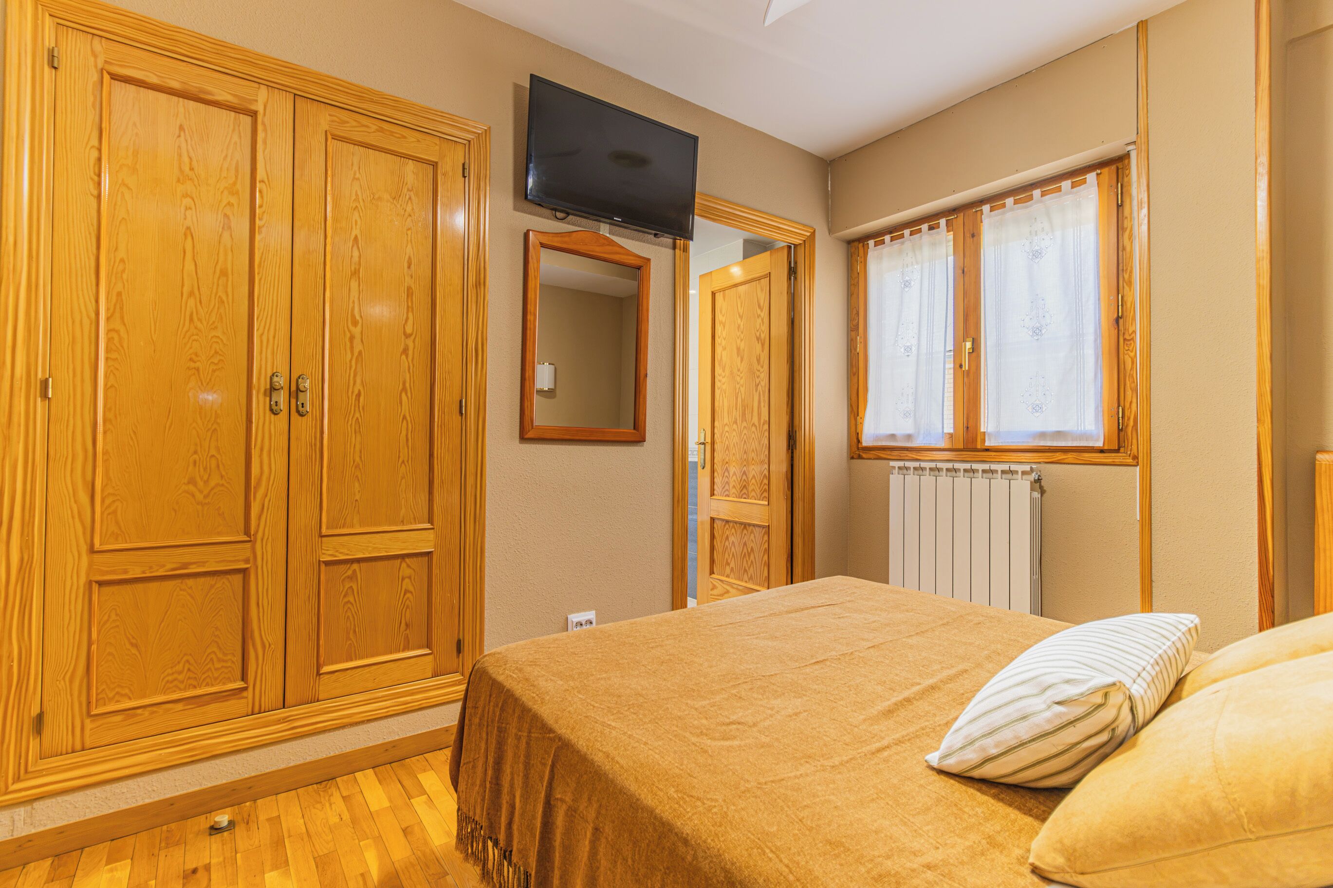 Een slaapkamer met een bed, een televisie, houten kasten, een raam met gordijnen en een radiator.