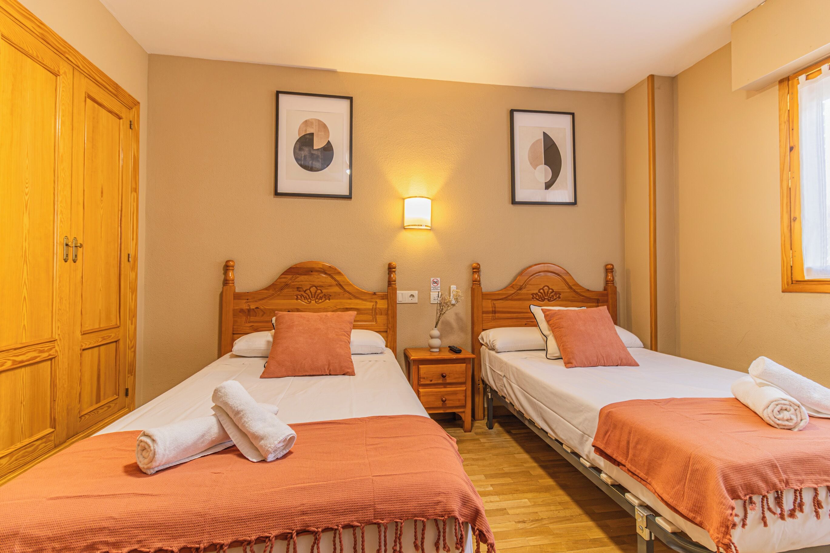 Habitació d'hotel amb dos llits, ambures de fusta, una petita taula de nit, una làmpada penjada a la paret i quadres amb marc.