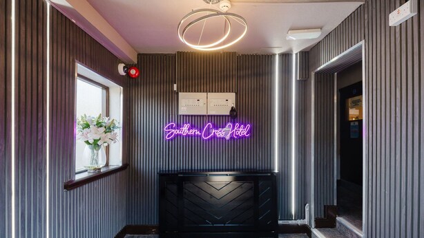 A hotel reception area with a neon sign, a reception desk, a window with flowers, and a modern light fixture. - Comfortable King Room with Private Bathroom (Watford)
