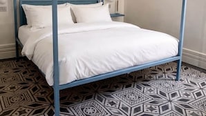 A four-poster bed with white bedding, a geometric patterned floor, and a plain wall with a light switch.