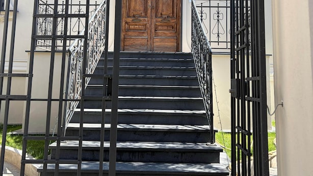 A set of black metal stairs leading to a wooden double door with a decorative emblem above it, flanked by black metal railings and wall-mounted light fixtures.