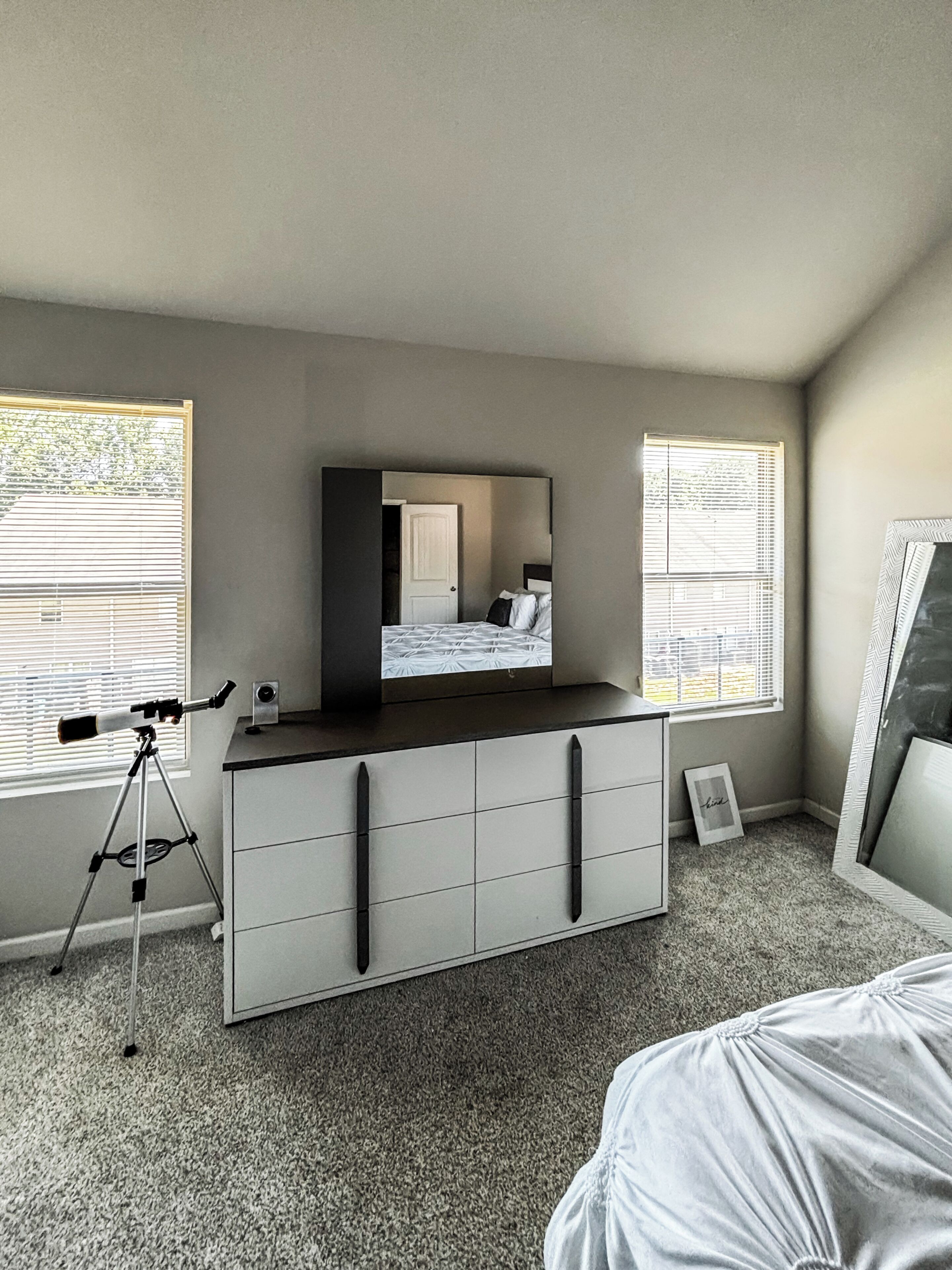 Une chambre à coucher comprenant un lit, une commode, un télescope et deux fenêtres équipées de stores.