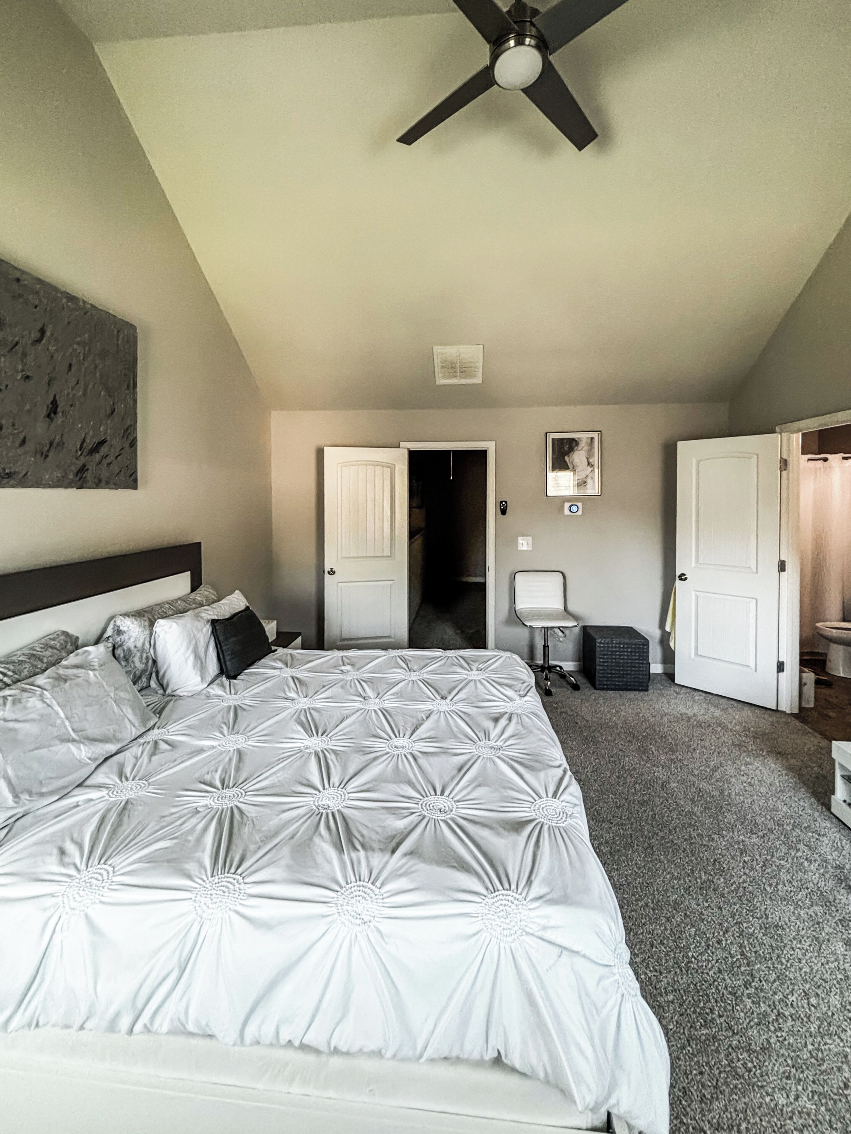 Un dormitorio con una cama grande, un ventilador de techo, una puerta que da a otra habitación y una mesita con una silla.