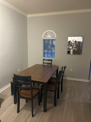 A dining area with a wooden table and chairs, a window with blinds, and a kitchen area with a refrigerator.