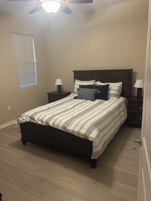 A bedroom with a bed, two nightstands, a ceiling fan, a window with blinds, and a door.