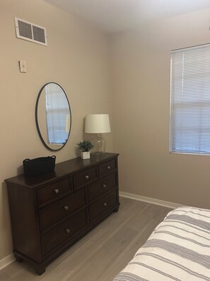 A bedroom with a dresser, a mirror, a lamp, and a window with blinds.