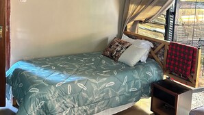 Una cama individual con una colcha estampada, una mesita de noche con un mantel a cuadros rojo y negro, y una mosquitera colgante.