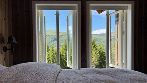 Ein Schlafzimmer mit einem großen Bett, zwei offenen Fenstern und Blick auf eine bewaldete Landschaft.