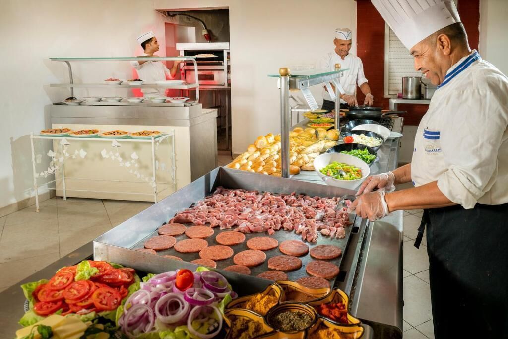 Un área de cocina con chefs preparando comida, una parrilla con carne y diversos alimentos en exhibición.