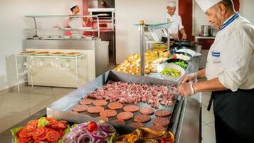 Un área de cocina con chefs preparando comida, una parrilla con carne y diversos alimentos en exhibición.