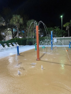 Uma área de recreação aquática com vários jatos d'água e uma piscina com espreguiçadeiras.