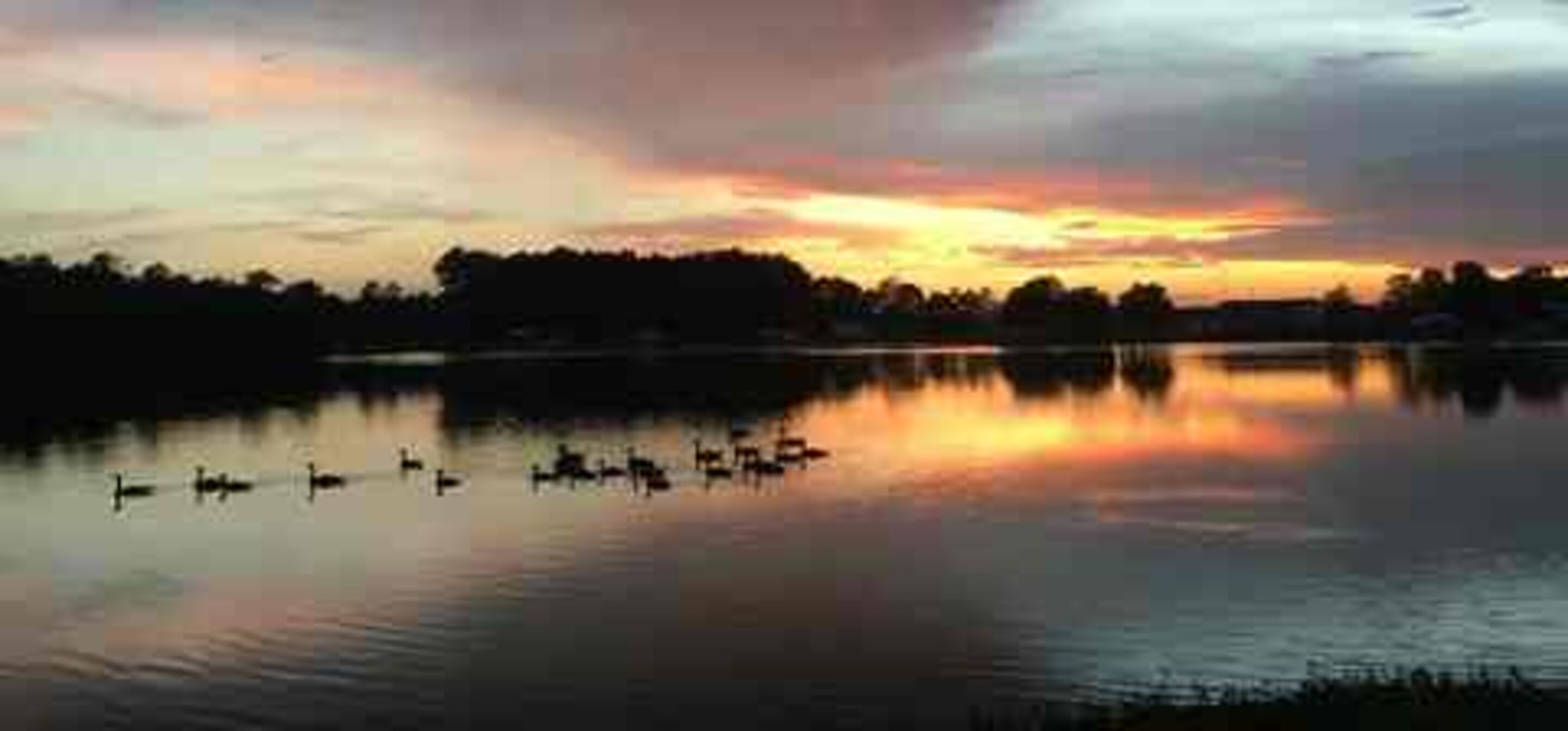 A calm lake at sunset with reflections of the sky and silhouettes of trees and boats.