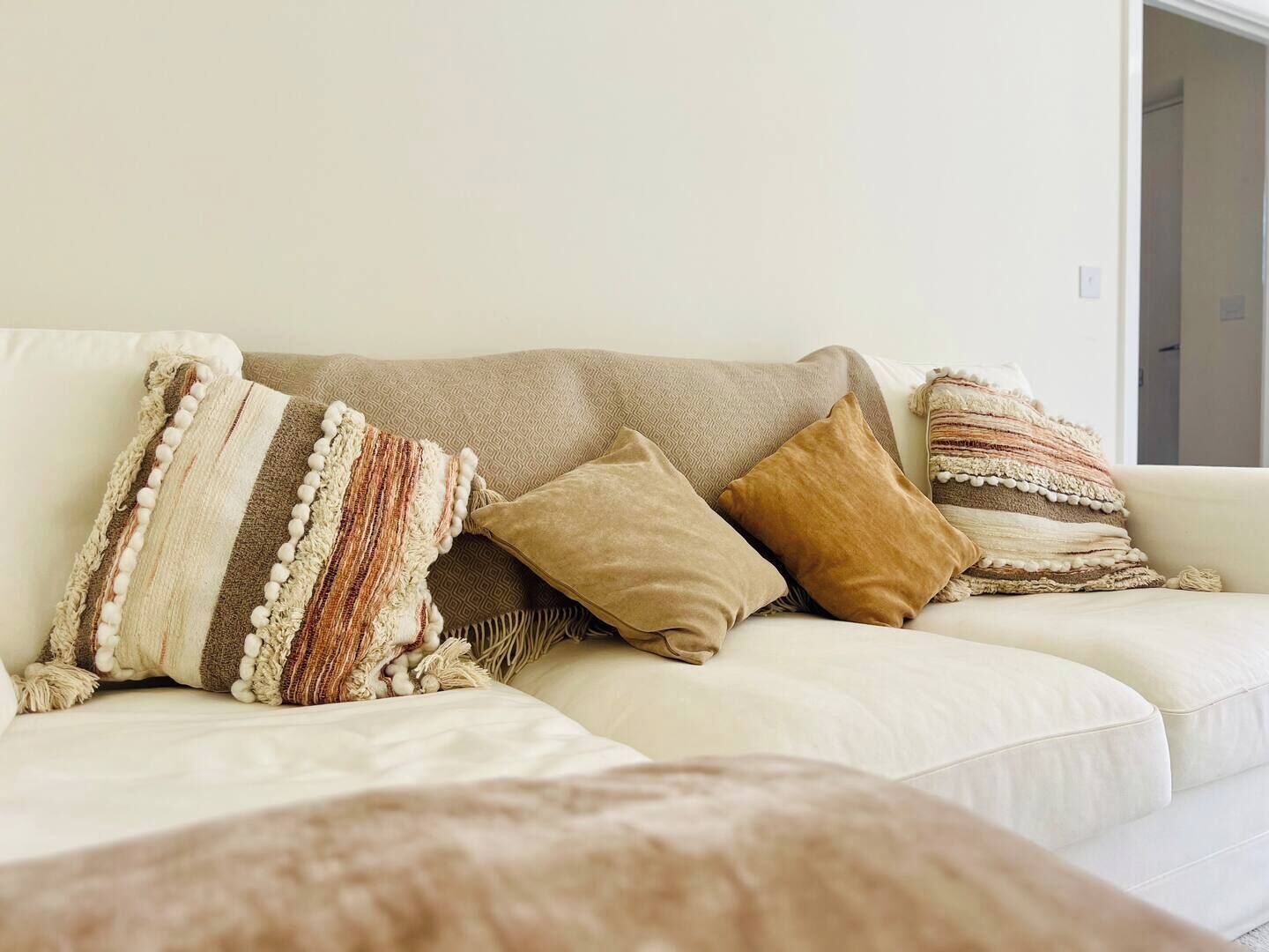 En beige soffa med tre kuddar, varav en randig och två enfärgade, i ett rum med en enkel vägg och en dörr i bakgrunden.