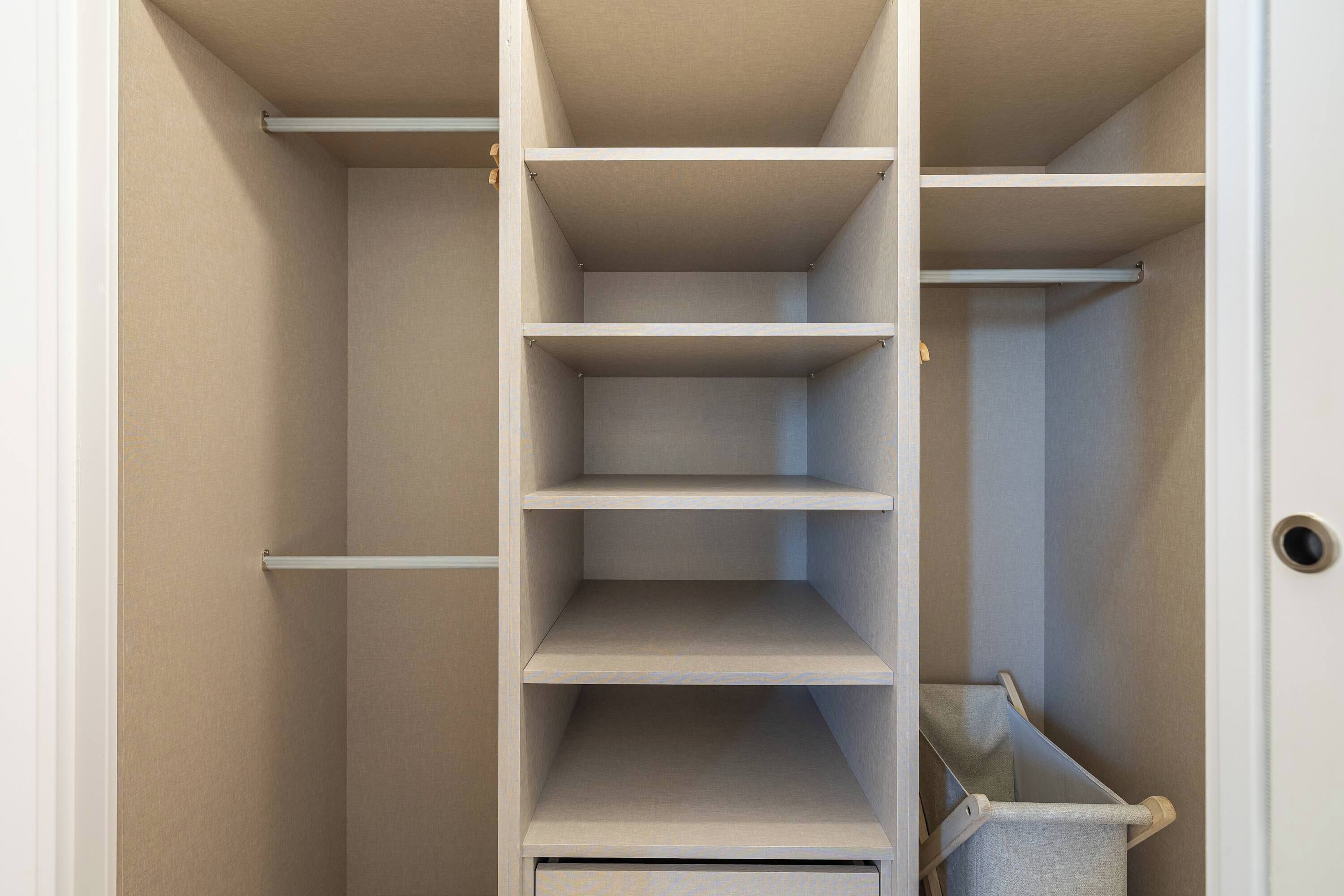 Ein Schrank mit mehreren Einlegeböden und Kleiderstange, einer Tür mit Griff sowie einem kleinen Mülleimer.