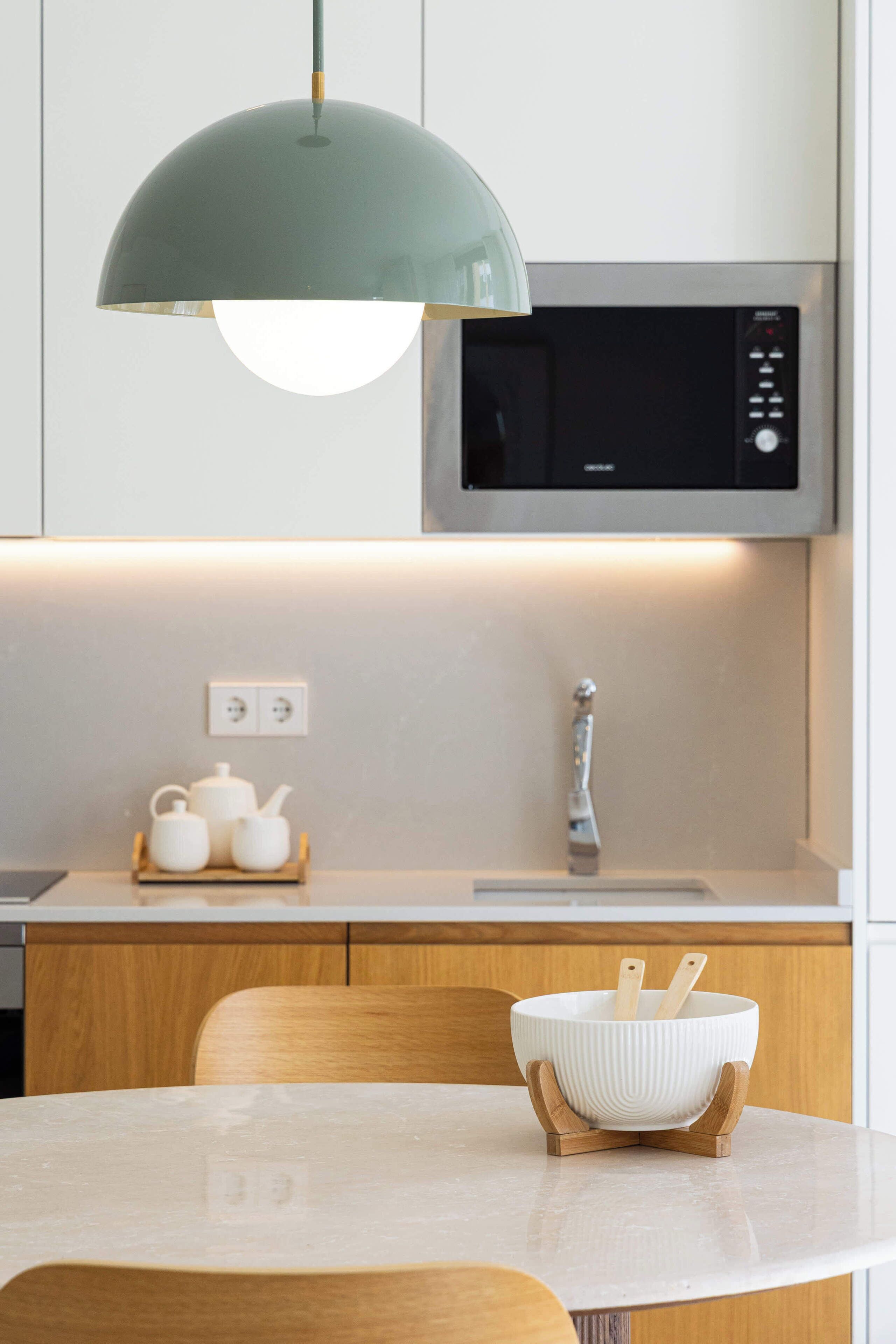 Una cocina moderna con encimera blanca, un cuenco de madera sobre un soporte, un microondas arriba y una lámpara colgante.