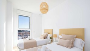Een hotelkamer met twee bedden, een groot raam met uitzicht op de stad, een hangende rotanlamp en een nachtkastje met een lamp.