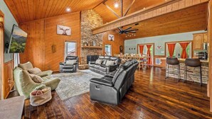 A spacious living room with a fireplace, leather sofas, a dining area, and a kitchen with stone accents. - 2 Cabins in One- Mtn Views - Hot Tub - Fenced Yard (Pigeon Forge)