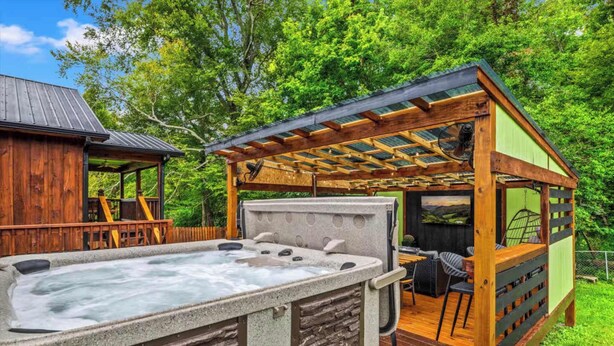 A hot tub with stone surround, wooden deck, and a covered seating area with chairs and a table. - 2 Cabins in One- Mtn Views - Hot Tub - Fenced Yard (Pigeon Forge)