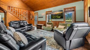 A spacious living room with leather sofas, a patterned rug, and a large TV screen displaying a scenic landscape. - 2 Cabins in One- Mtn Views - Hot Tub - Fenced Yard (Pigeon Forge)