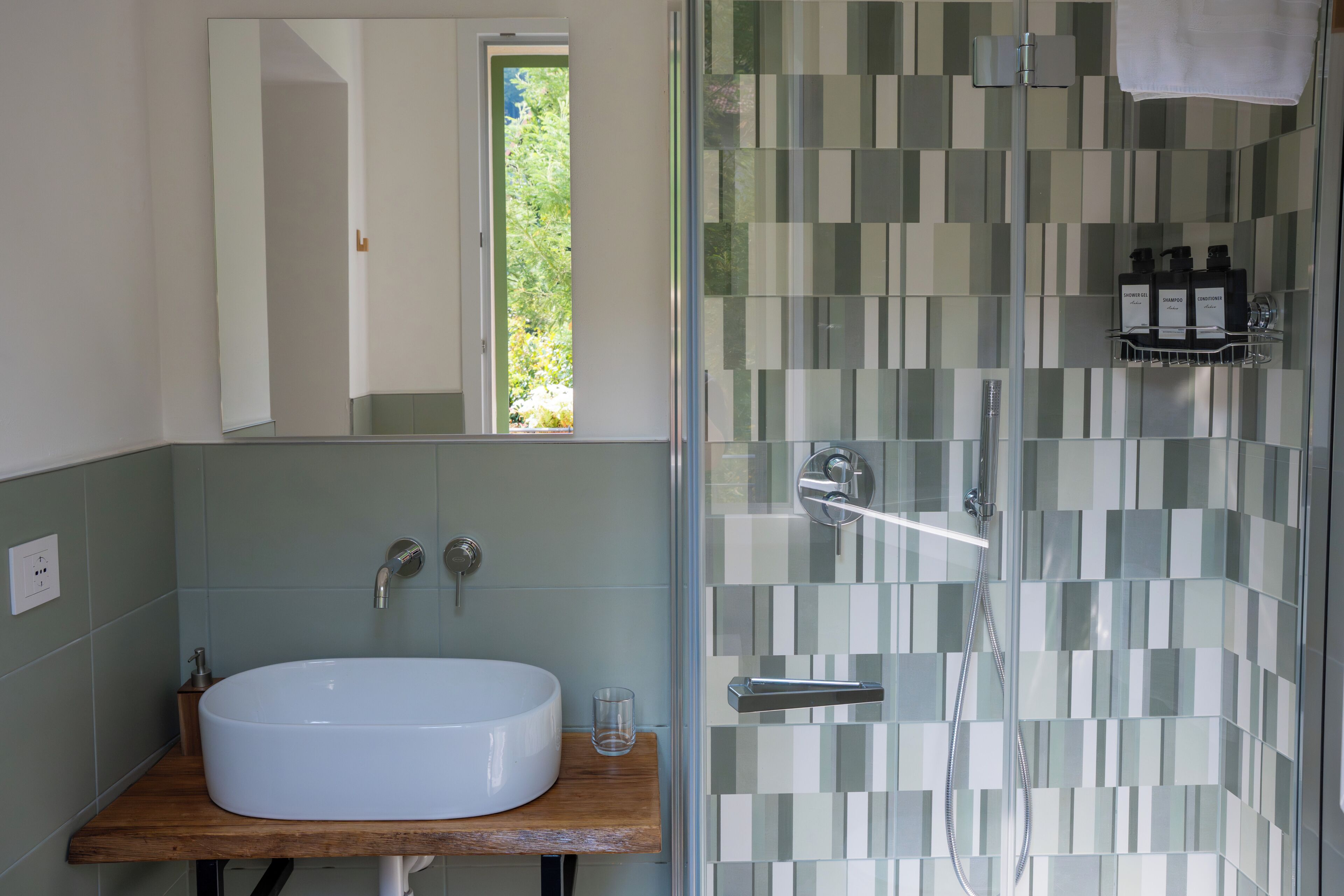 A modern bathroom with a freestanding bathtub, a glass shower enclosure, and a mirror reflecting a green outdoor view.