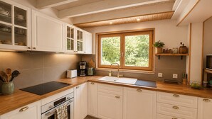 Une cuisine avec des meubles blancs, un plan de travail en bois, un four encastré, un évier et une fenêtre donnant sur un espace verdoyant.