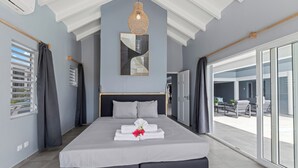 Une chambre moderne avec un grand lit, un ventilateur de plafond, un miroir fixé au mur et une porte-fenêtre coulissante donnant sur un espace extérieur.