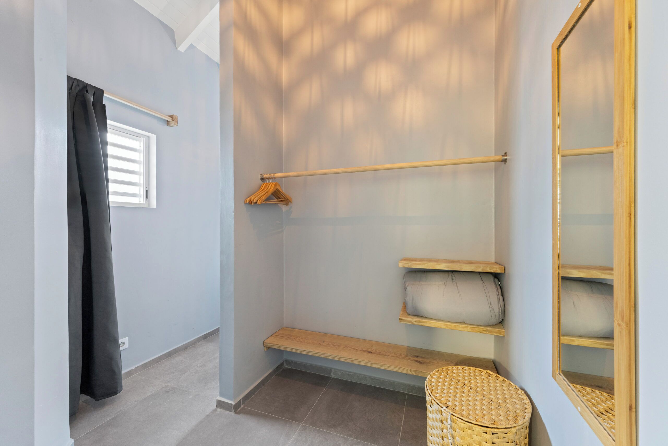 A room with a wooden bench, a hanging coat rack, a wicker basket, and a window with blinds.