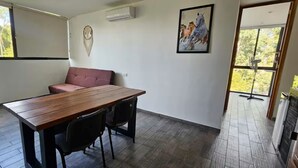 Una habitación con una mesa de madera, dos sillas, un sofá rosa, un aire acondicionado instalado en la pared y un cuadro enmarcado de caballos.