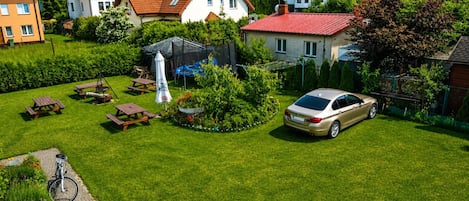 Ein Wohngebiet mit Häusern, einem Auto und einem Garten mit Picknicktischen und einer Schaukel.