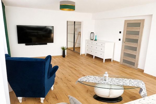 A living room with a blue chair, a glass coffee table, a white dresser, and a TV mounted on the wall.